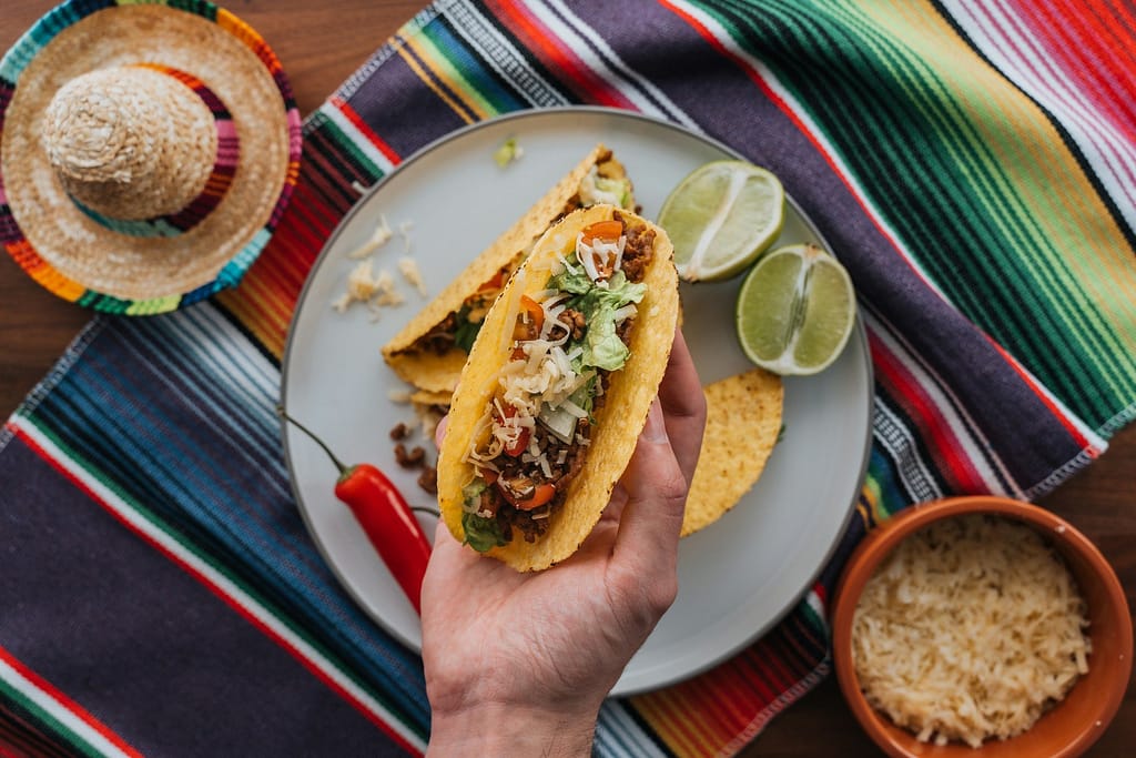 Hand holding a carne asada taco with lettuce, tomatoes, and cheese above a plate with lime wedges and tacos, on a colorful Mexican tablecloth